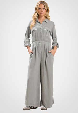 Kobieta ubrana w jasnoszary, zapinany na guziki kombinezon z szerokimi nogawkami, z podwiniętymi rękawami i dłońmi w kieszeniach, stojąca na jednolitym tle.
