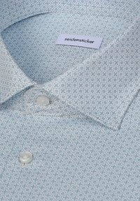 Chemise à manches longues pliée bleu clair à motifs avec des boutons blancs et une étiquette de marque "seidensticker" visible à l'intérieur du col.