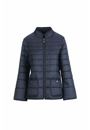 Woman wearing a navy quilted jacket with front pockets, white high-waist wide-leg pants, and beige pointed shoes, standing with one hand in pocket.