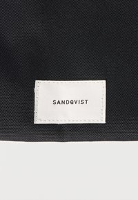 Čierna textúrovaná látková etiketa s bielym prešitím. Na štítku je čiernym veľkým písmom nápis "SANDQVIST". Jednoduchý dizajn bez vzorov alebo zdobení.