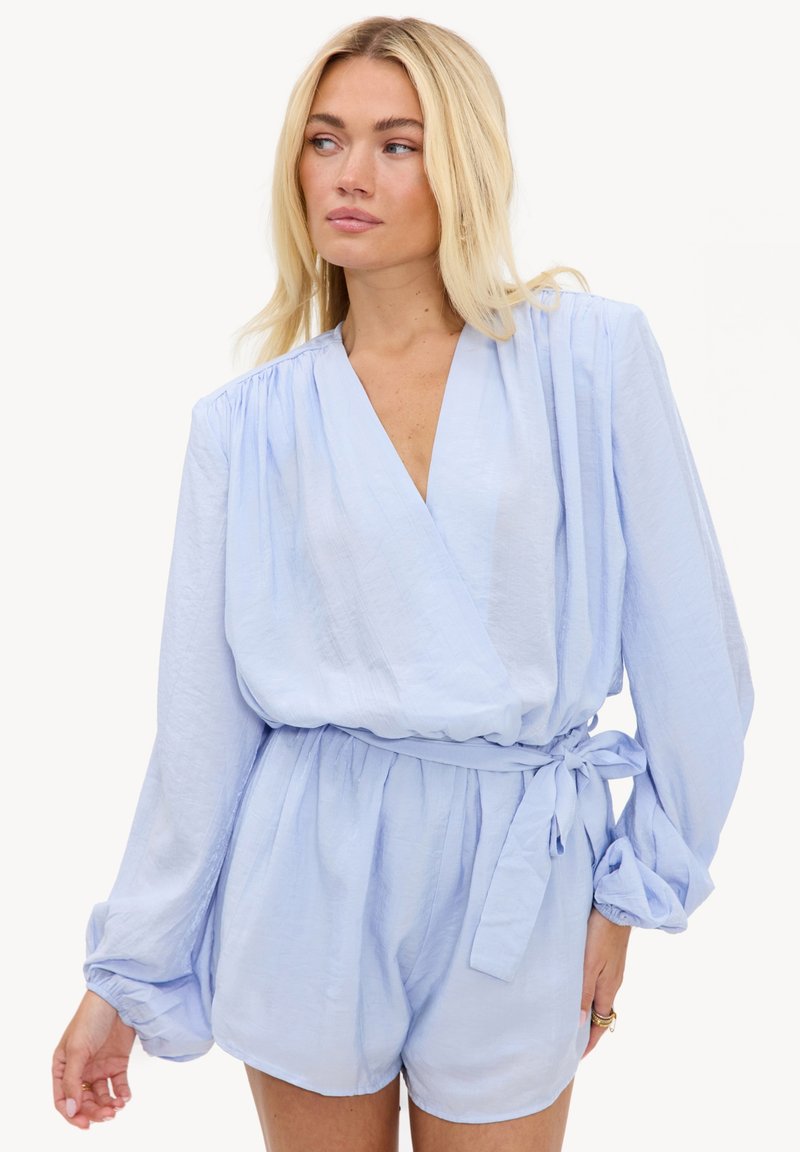 Femme aux longs cheveux blonds portant une combinaison cache-cœur à manches longues bleu clair avec une taille nouée, regardant sur le côté devant un fond uni.