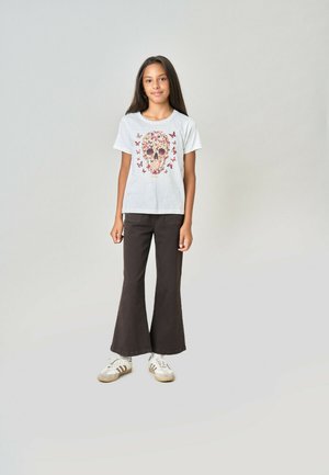 Fille debout portant un t-shirt blanc avec un motif papillon-crâne, un pantalon noir évasé et des baskets blanches sur fond gris clair.