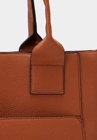 Close-up van een bruine, getextureerde leren handtas met zichtbare handvatbevestiging en gedetailleerd stiksel.