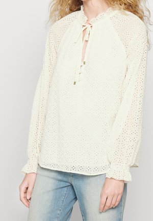 Vrouw met een crèmekleurige blouse van oogjeskant met lange geplooide mouwen en een striksluiting bij de hals, gecombineerd met lichtblauwe spijkerbroek.