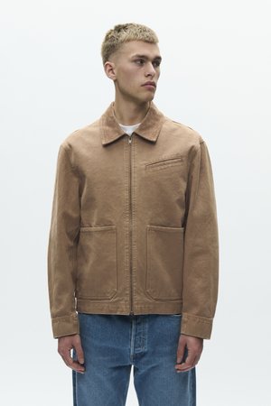 Mladý muž v pískovohnedej bunde na zips s golierom a dvomi prednými vreckami, v modrých džínsoch, stojaci pred jednoduchým pozadím.