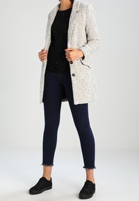 Abrigo texturizado gris claro con cuello de muesca, dos botones, llevado sobre una blusa negra con adornos, jeans oscuros y zapatillas negras.