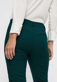 Ciemnozielone dopasowane spodnie o gładkiej fakturze, z tylnymi kieszeniami, w połączeniu z białą bluzką. Widoczna ręka odpoczywająca na biodrze.