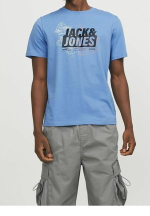 Mężczyzna w jasnoniebieskiej koszulce z logo Jack & Jones i szarych szortach typu cargo na jednolitym tle, ręce swobodnie opuszczone wzdłuż ciała.