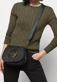 Svart skinnväska med texturerad yta, rund gulddetalj och justerbar rem, bärs med en grön kabelstickad tröja.