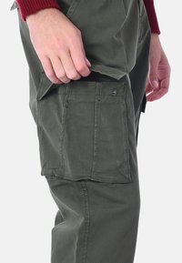 Pantaloni cargo verde oliva con tasca laterale, realizzati in un tessuto resistente. Presenta dettagli di cucitura e un robusto hardware con bottoni.
