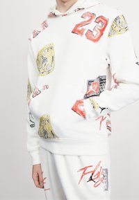Sudadera blanca con estampados gráficos variados, incluidos números y logotipos en rojo, amarillo y azul. Presenta un bolsillo frontal y capucha con cordón.