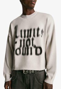 Svetlošedý skrátený sveter s okrúhlym výstrihom, s čiernym gradientovým potlačou slova "Limit" a voľným strihom. Nosený s tmavými džínsami.