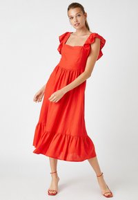 Vestido rojo sin mangas con escote cuadrado, acentos de hombros con volantes y un dobladillo en capas. Hecho de una tela ligera con una silueta fluida.