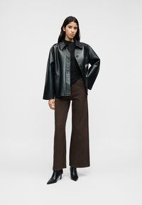 Veste en cuir noir à boutons sur le devant, portée par-dessus un haut noir ajusté, associée à un pantalon large marron et des bottines noires à talons pointues.