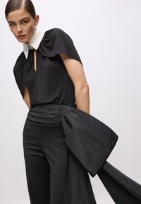 Schwarze Bluse mit Capeärmel und einem weißen Kragen, kombiniert mit maßgeschneiderten schwarzen Hosen mit einem gefalteten Bund und einem gebundenen Detail.