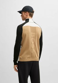 Strickpullover in Beige, Schwarz und Weiß. Mit hohem Kragen, enganliegenden Ärmeln und großem 'BOSS'-Logo auf dem Rücken in tonalem Relief.