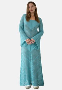 Turkusowa dzianinowa sukienka z długimi rękawami i fasonem dzwonowym, wzorzysta w zygzaki i dopasowana do kostek.