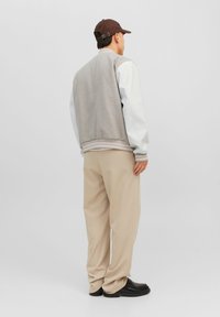 Man in beige broek, grijs-witte varsityjack, bruine pet en zwarte schoenen die met zijn rug naar de camera staat tegen een effen lichte achtergrond.
