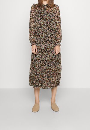 Vrouw staand, gekleed in een zwarte midi-jurk met lange mouwen en bloemenprint en beige geweven instapschoenen tegen een effen witte achtergrond.