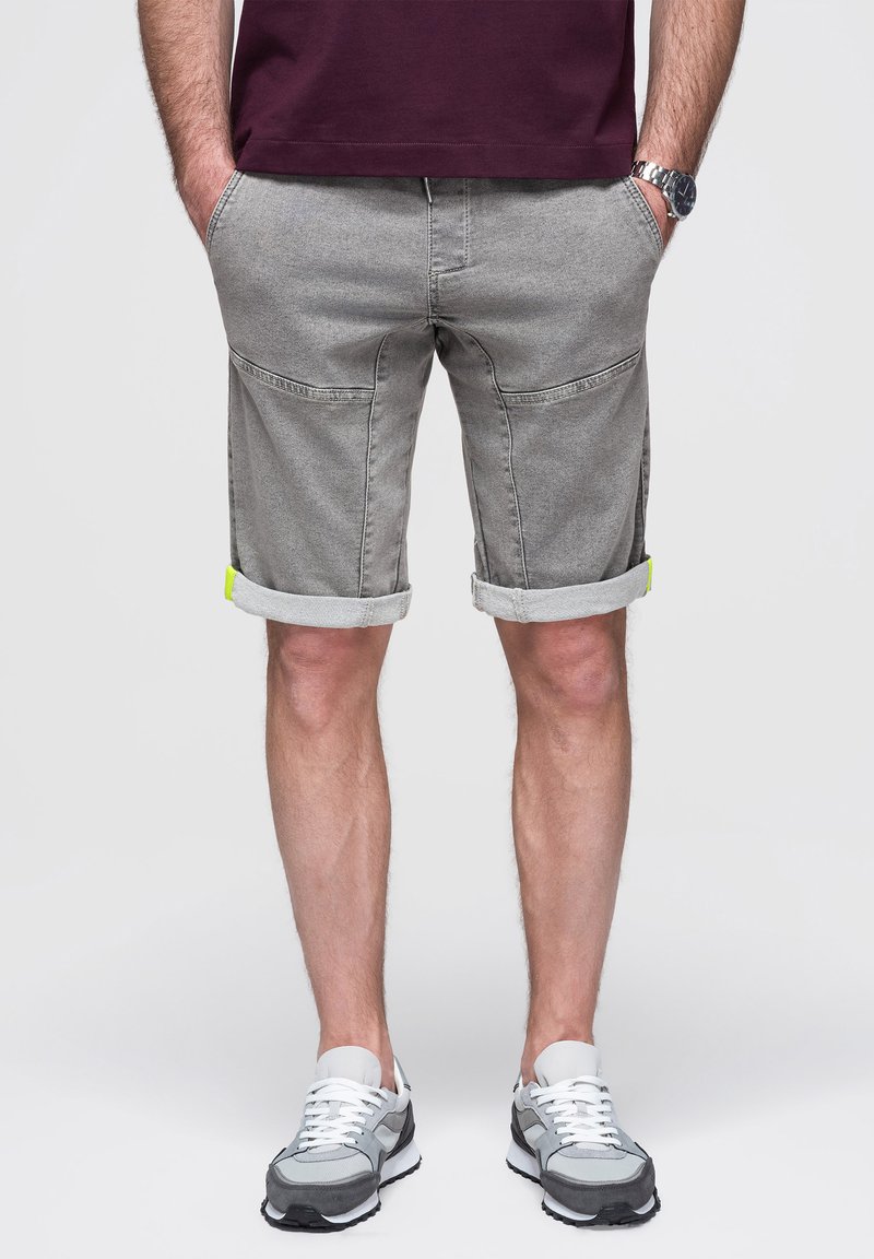 Homme portant un short gris plié, des baskets blanches et grises, un t-shirt bordeaux et une montre argentée, debout les mains dans les poches.