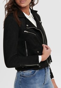 Czarna zamszowa kurtka motocyklowa z zamkami błyskawicznymi, zatrzaskami i dopasowanym fasonem. Noszona na białej koszuli i niebieskich dżinsach.