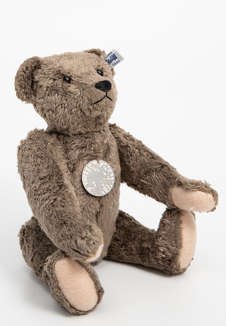 Brauner Plüschbär mit gestickten Details, weicher Textur, sitzender Haltung und einem metallischen Anhänger, der am Hals befestigt ist.