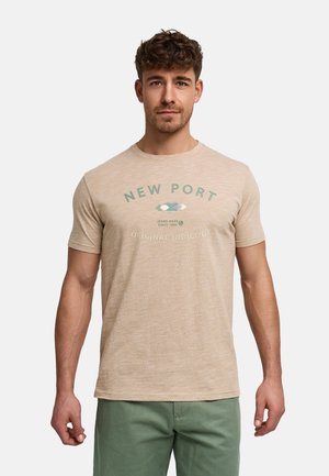 Mann mit kurzen lockigen Haaren, der ein beiges T-Shirt mit dem Aufdruck "New Port" und grüne Hose trägt, steht vor einem schlichten hellen Hintergrund.