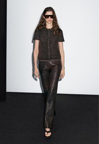 Frau, die auf dem Laufsteg geht und dunkle Sonnenbrillen, eine flauschige, geknöpfte Weste, ein schwarzes kurzärmliges Hemd, schwarze Lederhosen und Sandalen mit offenen Zehen trägt.