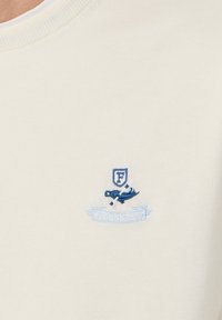 Tela color crema con un emblema bordado en azul que presenta un escudo, remos cruzados y una pancarta que dice "FACONNABLE" debajo.