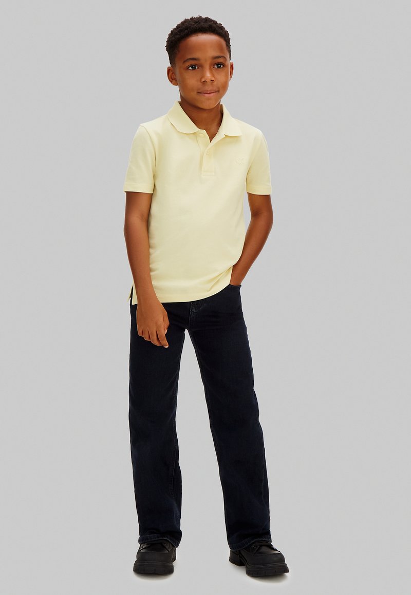 Garçon debout, une main dans la poche, portant un polo jaune clair, un pantalon noir et des chaussures noires, sur un fond gris uni.