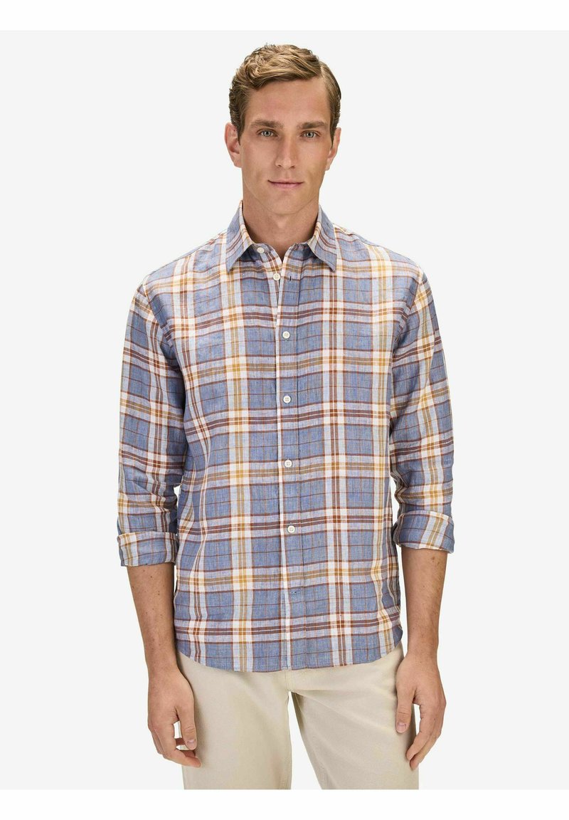 Homme portant une chemise à carreaux bleue, blanche et orange à manches longues retroussées jusqu'aux coudes, debout devant un fond clair uni.