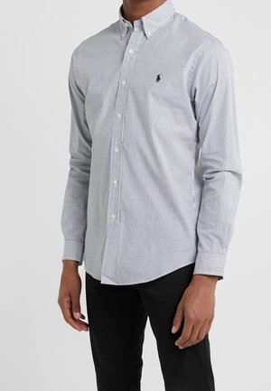 Homme portant une chemise blanche cintrée à manches longues avec de fines rayures verticales noires et un pantalon noir, debout devant un fond uni.
