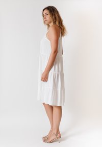 Abito maxi bianco a strati con design senza maniche, realizzato in tessuto leggero. Modello che indossa tacchi metallici con cinturini. Sfondo neutro.