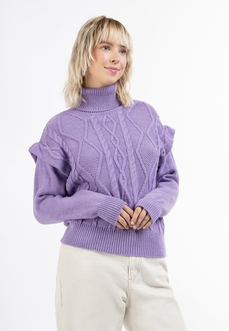 Pulover tricotat cu guler înalt de culoare liliachiu, având un design texturat cu cabluri, manșete și tiv nervurat. Modelul poartă pantaloni cremuți cu o croială lejeră.
