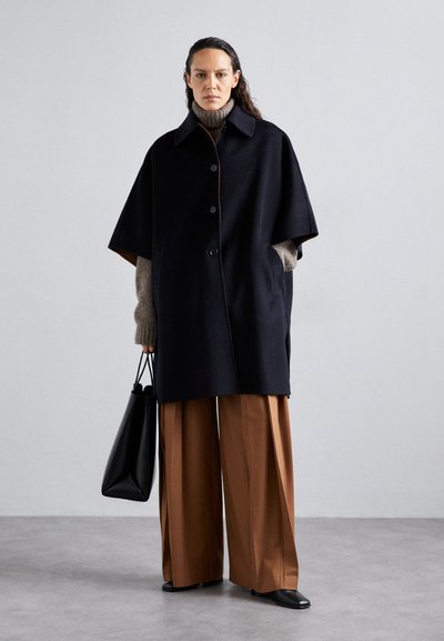 Cappotto oversize nero con maniche corte, chiusura a bottoni e tasche, indossato sopra un dolcevita beige. Abbina pantaloni larghi marroni e una borsa nera.