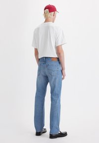 Lightweight white t-shirt, relaxed fit. Denim jeans in light blue, straight-leg design, visible back pockets, and leather patch detail. Black shoes.