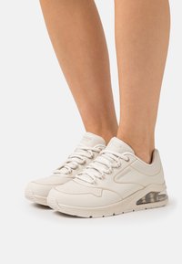 White sneakers with a textured pattern, rounded toe, and clear air cushioning in the heel. Lace-up design and "SKECHERS" logo visible.