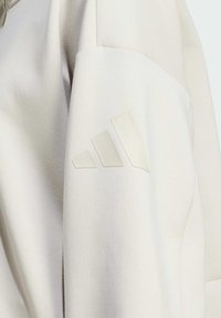 Svetlo béžová mikina s jemnou textúrou. Má zdvihnuté, tónové logo s tromi pruhmi na ľavom rukáve.