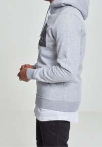 Felpa con cappuccio grigia in materiale morbido, con tasca frontale e dettaglio logo nero. Presenta orlo e polsini a costine, indossata sopra uno strato bianco.