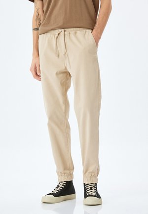 Beige bomuldsjoggers med elastisk talje og manchetter, der har sidelommer. Sammen med sorte høje sneakers.