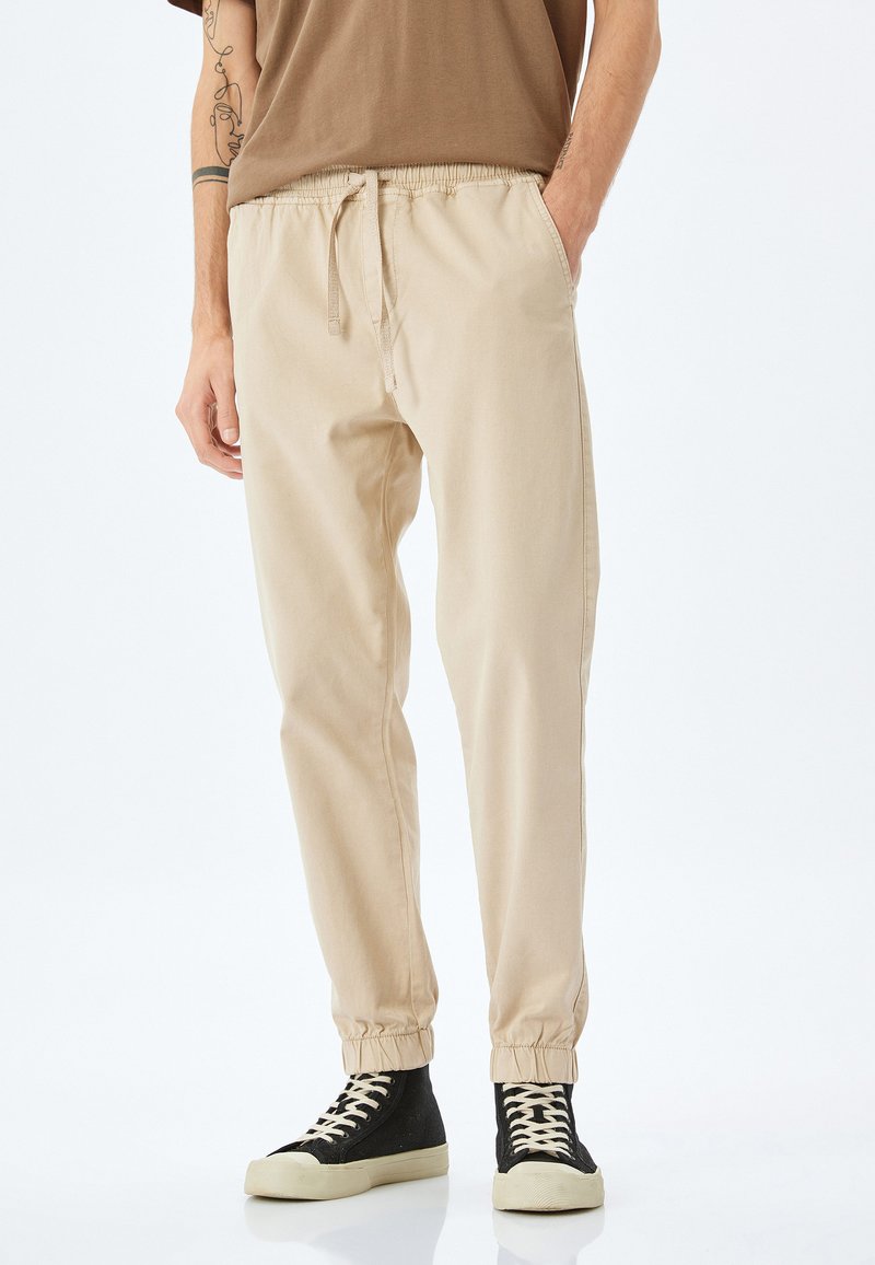 Beige katoenen jogpants met een elastische tailleband en boorden, met zijzakken. Gekoppeld aan zwarte hoge sneakers.