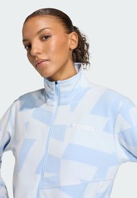 Fleeceová bunda v svetlomodrej farbe s geometrickými vzormi, vysokým golierom, plným zipsom a vyšitým logom TERREX na hrudi. Mäkká textúra.