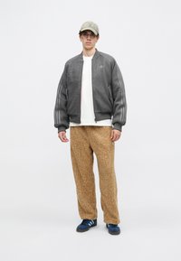 Chaqueta bomber gris con tela texturizada, camiseta blanca, pantalones marrón de felpa y zapatillas azules con tres rayas. El modelo lleva una gorra clara.