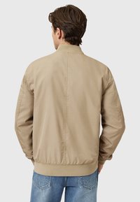 Chaqueta bomber beige con una textura suave, puños y dobladillo acanalados. Presenta un cuello levantado y un diseño sencillo en la parte posterior, combinada con jeans azules.