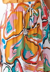 Detailní záběr barevné sukně s abstraktními květinovými vzory v oranžové, zelené, modré, růžové a červené barvě, s bílým šňůrkem v pase.
