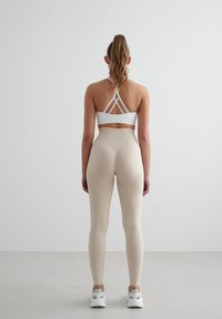 Witte sportbh met een crisscross-achterkant, gecombineerd met hoge beige leggings. Het model staat tegen een effen witte achtergrond.