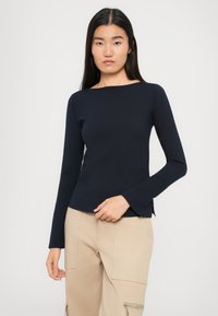 Tânără cu păr lung și închis la culoare, îmbrăcată într-un top albastru închis cu mânecă lungă și pantaloni cargo bej, pe un fundal simplu și deschis.
