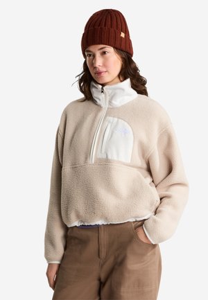 Kvinde iført en beige fleecejakke med en hvid brystlomme og en brun strikket hue, stående med den ene hånd i lommen.