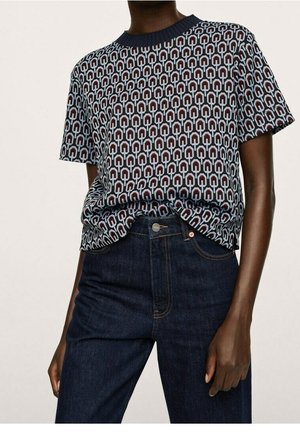 Osoba ubrana w krótki wzorzysty sweter z krótkim rękawem wpuszczony do wysokich, ciemnoniebieskich dżinsów, stojąca na jednolitym tle.