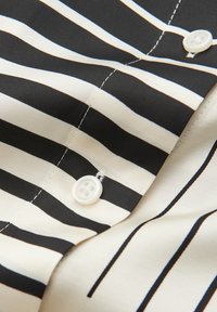 Gestreifter Stoff mit abwechselnden schwarzen und cremefarbenen Linien, versehen mit einem weißen Knopf und sichtbaren Nähten. Glatte Textur mit einem maßgeschneiderten Design.